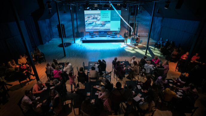 Kolme henkilöä lavalla ja yleisöä pöytien ääressä istumassa